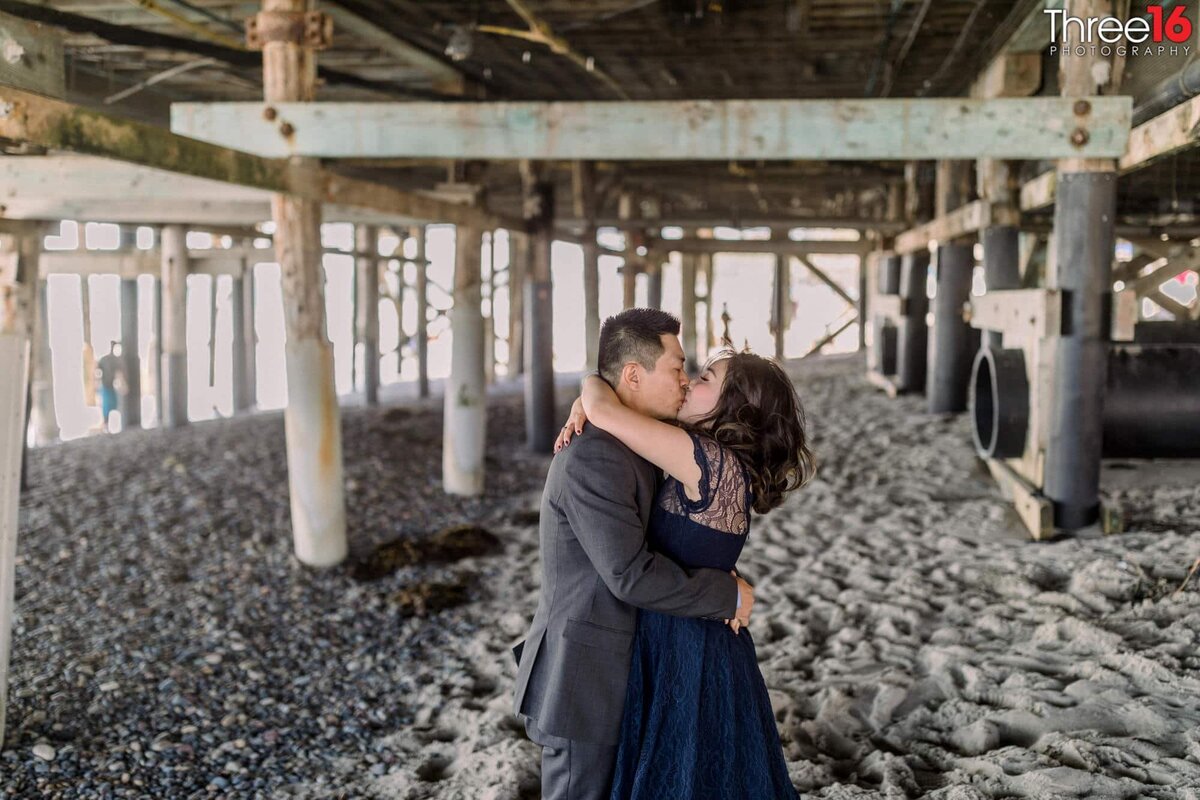 Engaged couple embrace with a big kiss while under the San Clemente Pier