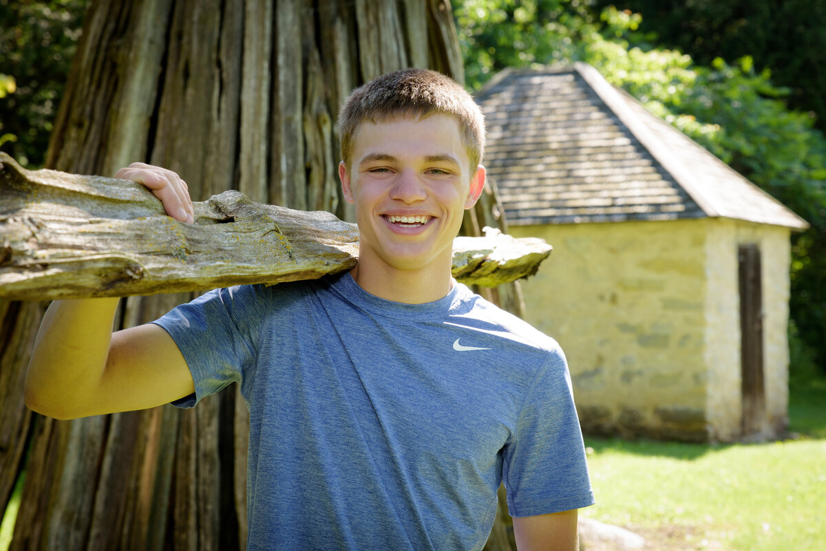 Luxemburg Casco high school senior boy wearing a short sleeve blue t-shirt and jeans holding wooden fence post by cabin at Devil's River Campground near Green Bay, Wisconsin.