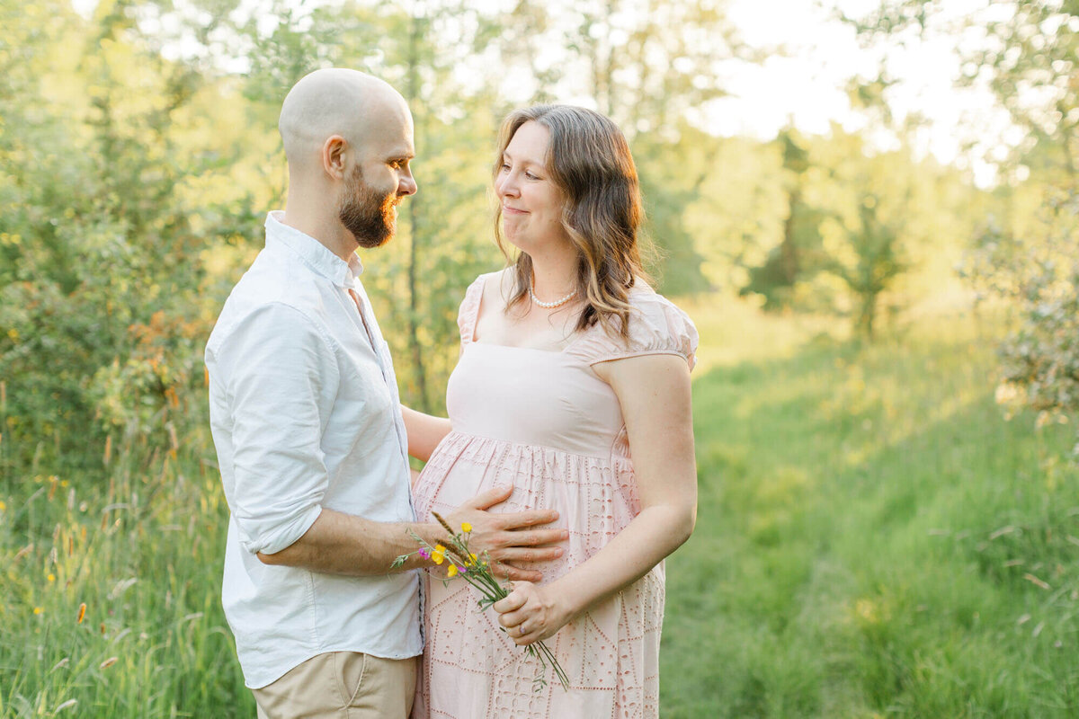 Pregnant mom in pink dress is holding some wildflowers and looking at dad who has his hands on her belly. Dad is dressed in a white button-down and khakis. They are smiling at each other. They are standing in a field of tall green grasses at sunset.