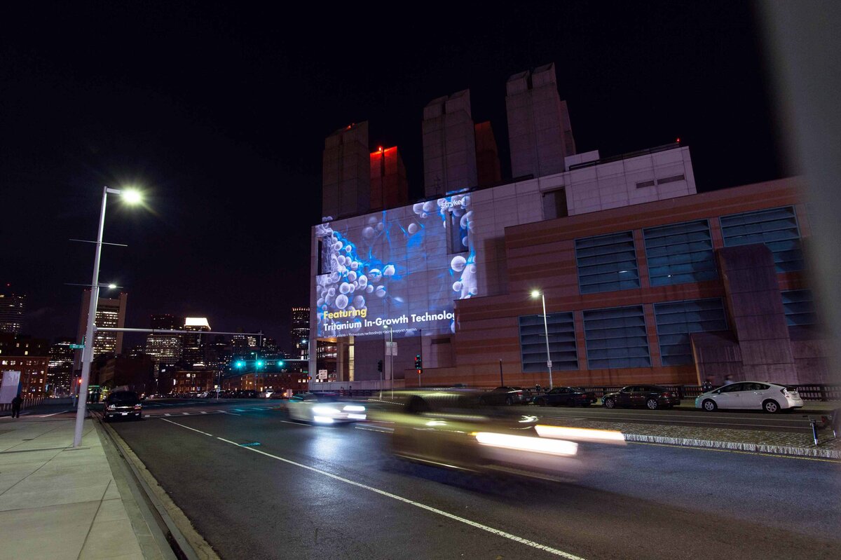 A project ad for event sponser stryker spinal implants is projected on a building at night with blurred cars driving down the street at conference