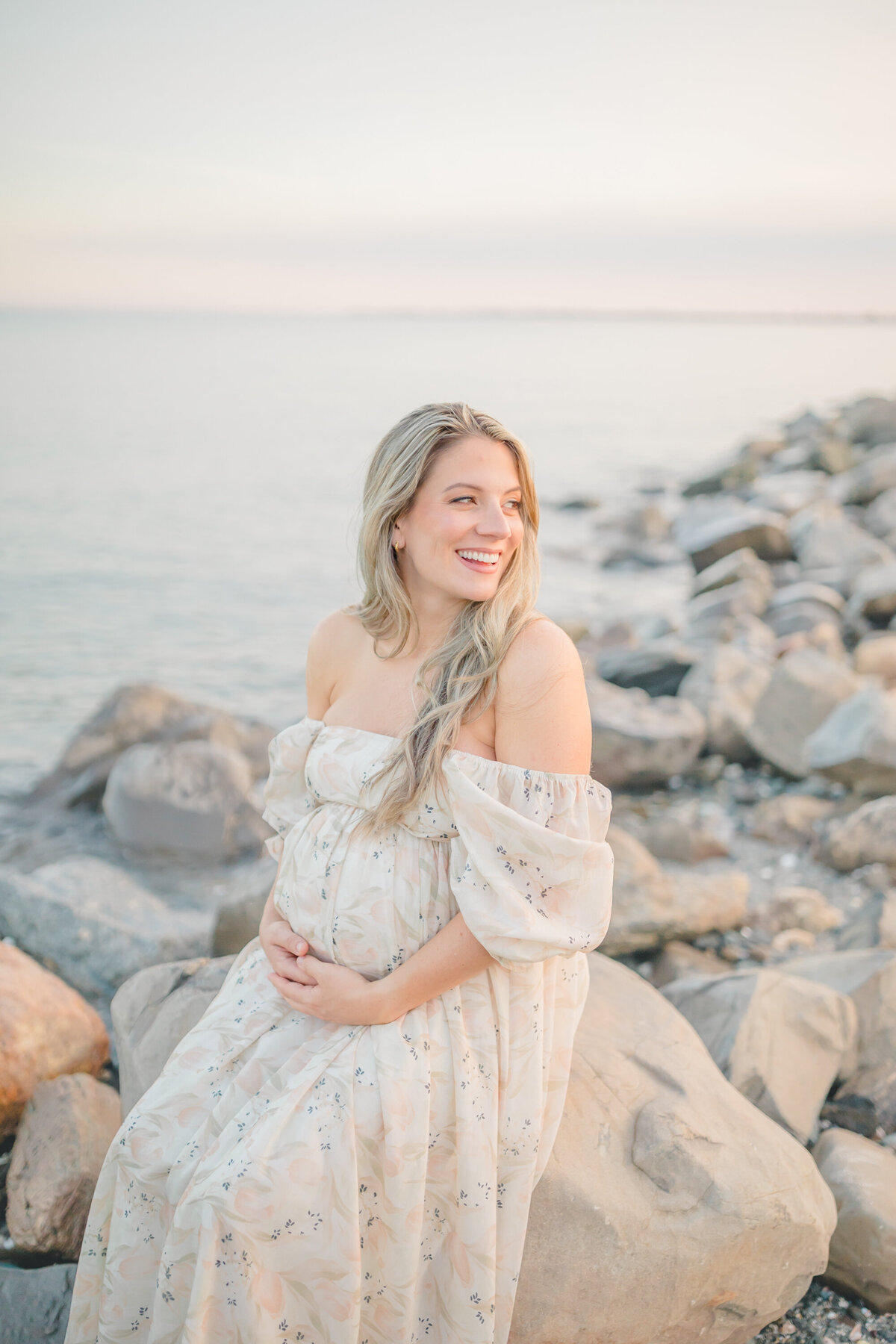 A pregnant woman in a flowing off-the-shoulder floral dress sits on rocks by the water during sunset, smiling and cradling her belly. Captured in soft, golden light by Westport photographer Oak & Lore Studio
