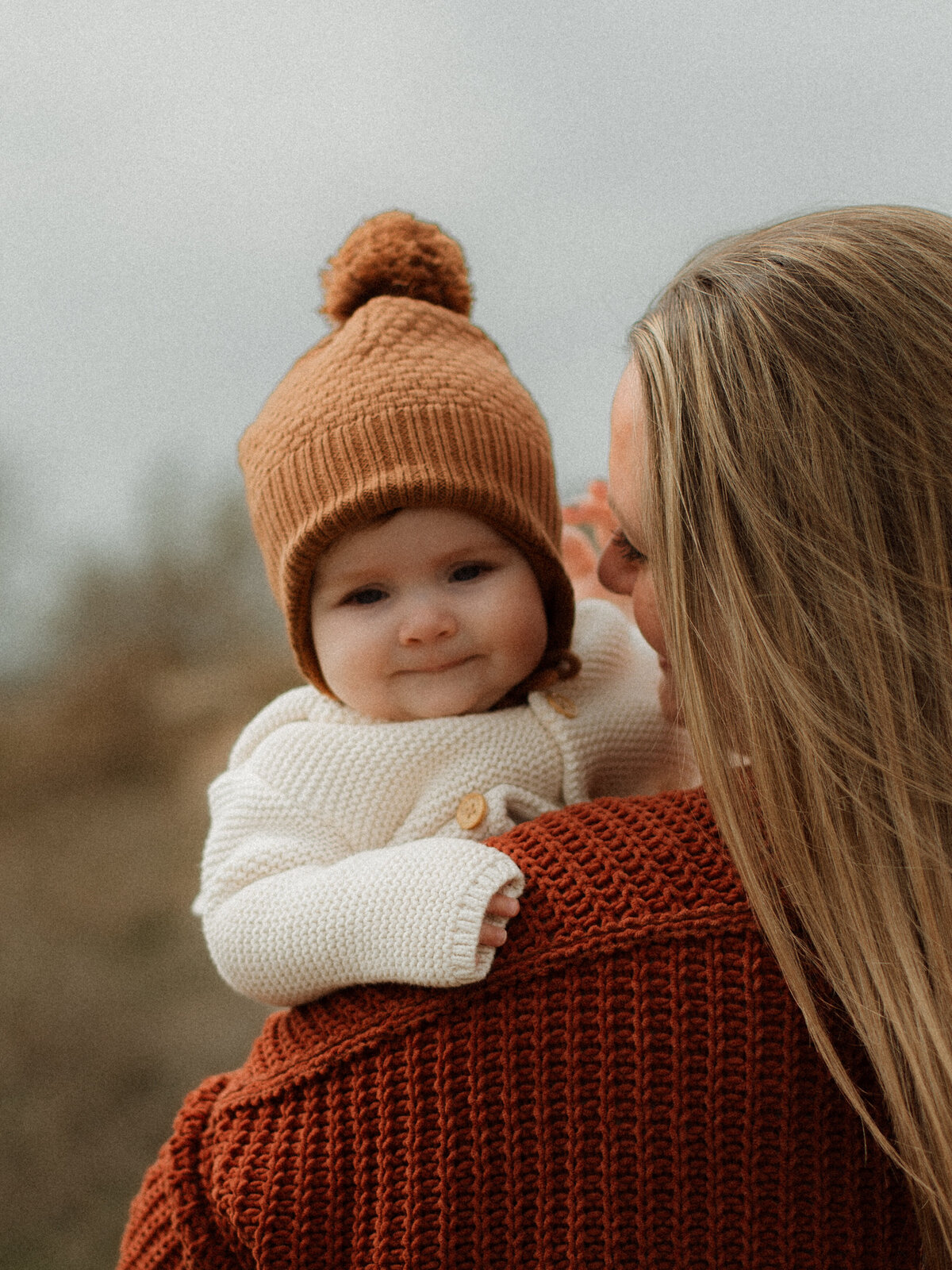 A baby in a knit cream sweater and brown beanie being carried by her mother, looking over her shoulder and smiling softly.