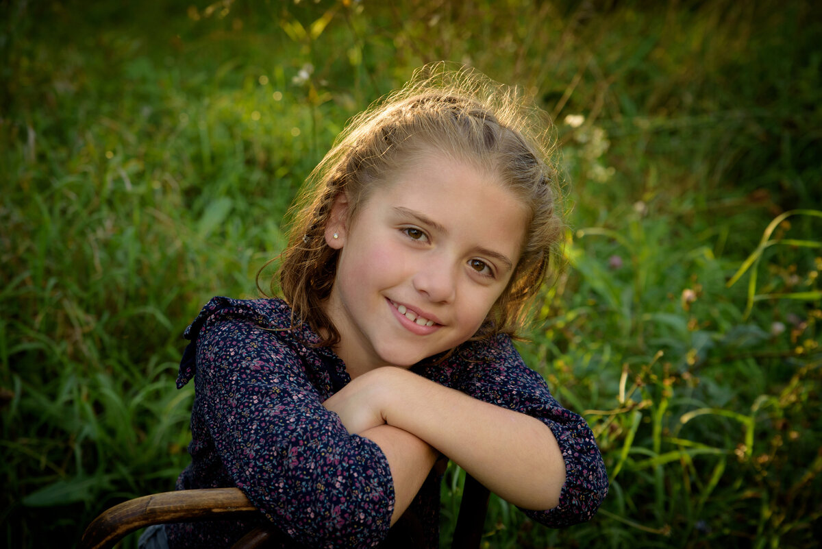 Young girl wearing a navy shirt and jeans sitting in a wooden chair in a long grassy field at Fonferek Glen County Park near Green Bay, Wisconsin