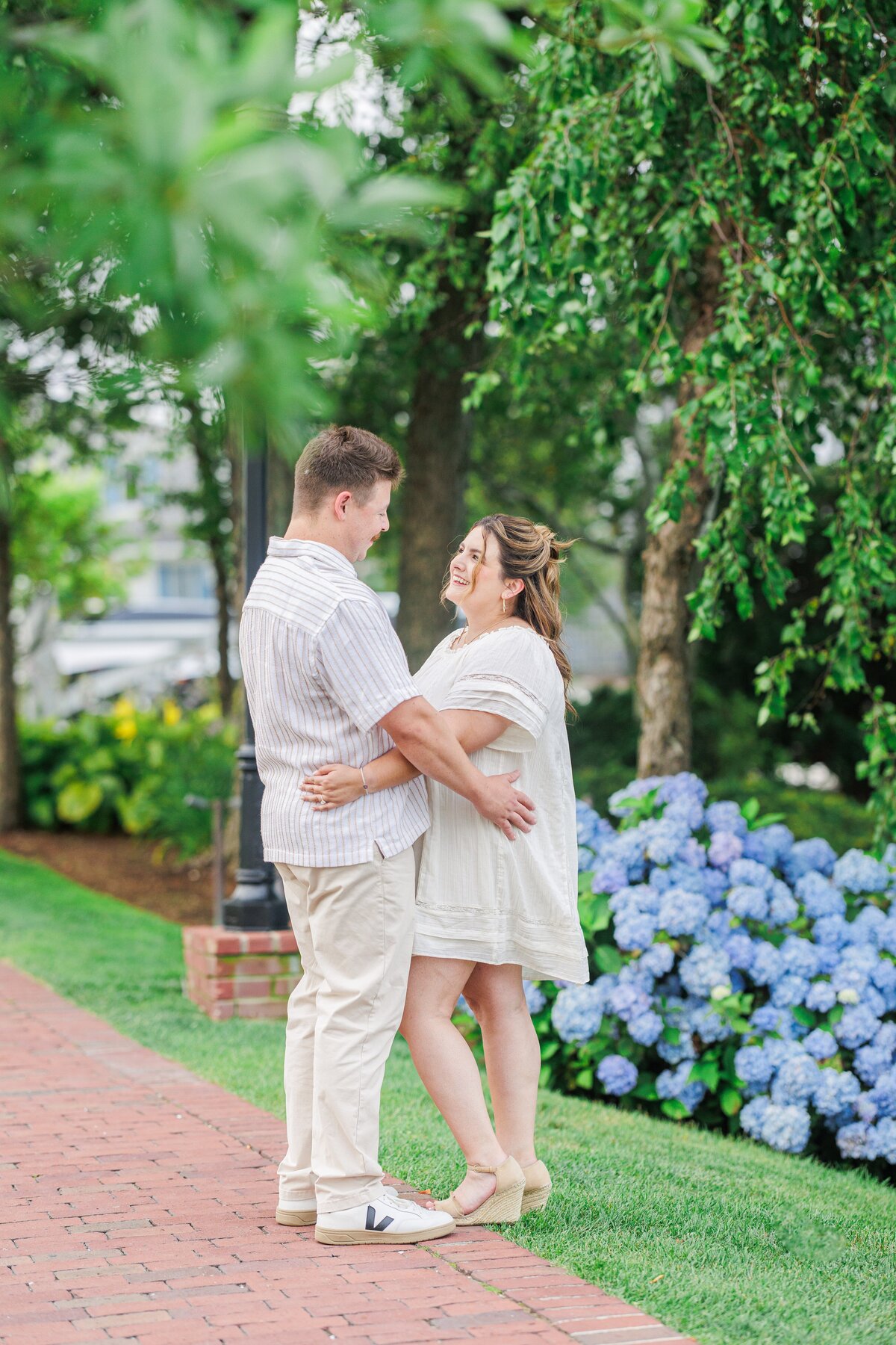 Couple standing face-to-face on a brick walkway, smiling and holding each other, with a row of blue hydrangeas and lush greenery in the background at Chatham Bars Inn in Chatham, Massachusetts — representing the portfolio of a Cape Cod engagement photographer.
