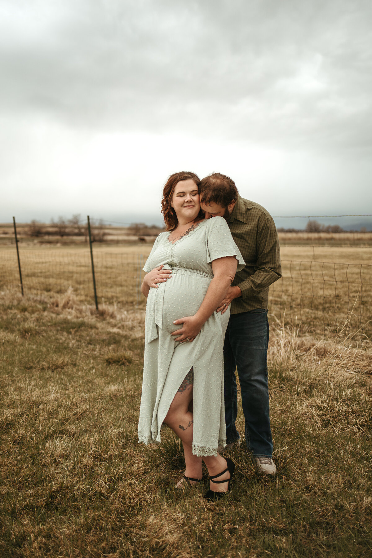 expectant dad kissing expectant moms shoulder while shes looking towards him smiling while holding twin baby girl belly in longmont colorado during maternity session with alexis adkins photography