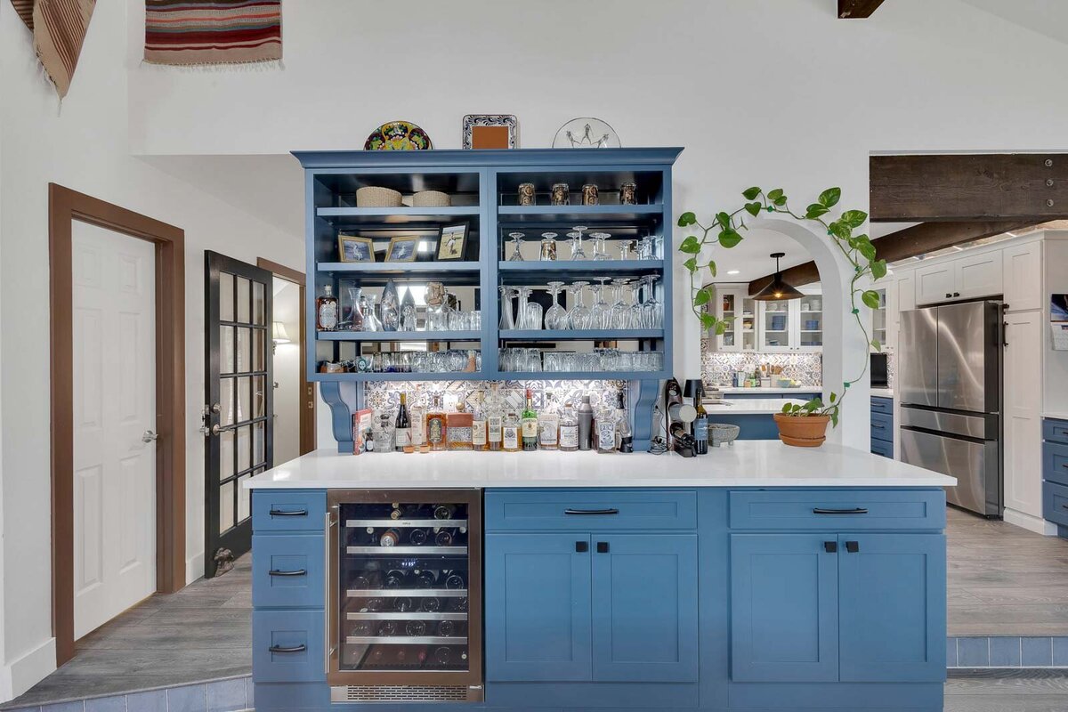 A wine bar with blue shelfs and cabinets with an arching window looking into the kitchen. 