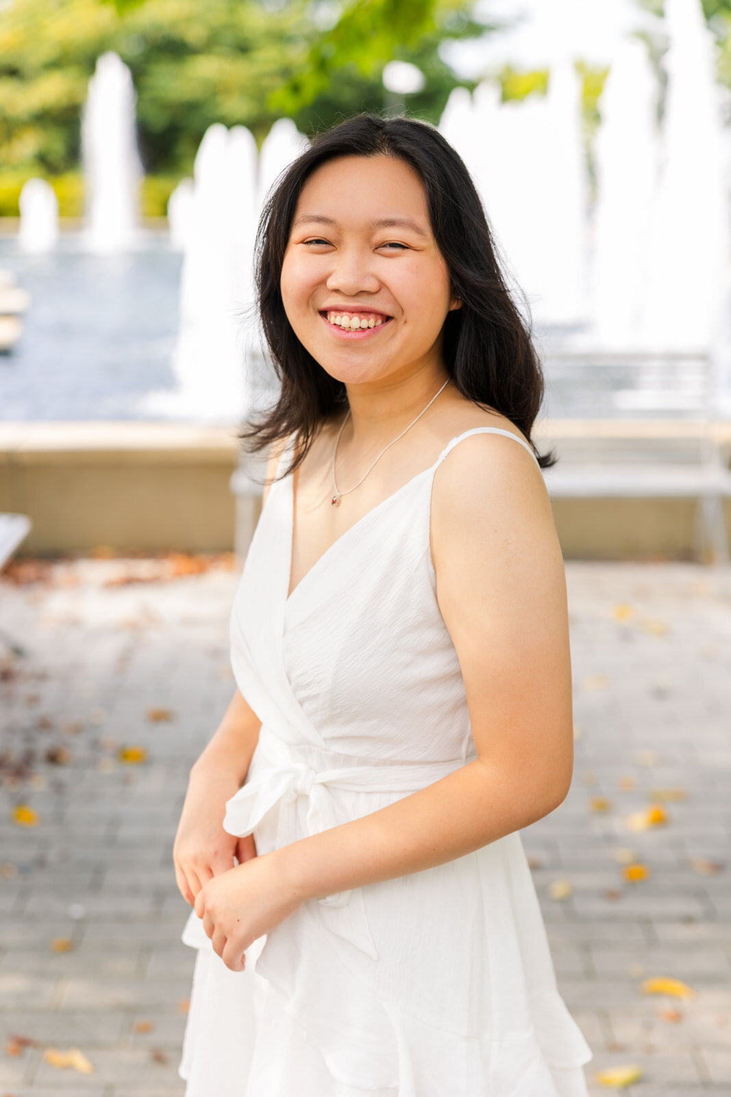 young woman wear a white sleeveless dress hands together and looking sideway at the camera in front of a fountain during photos