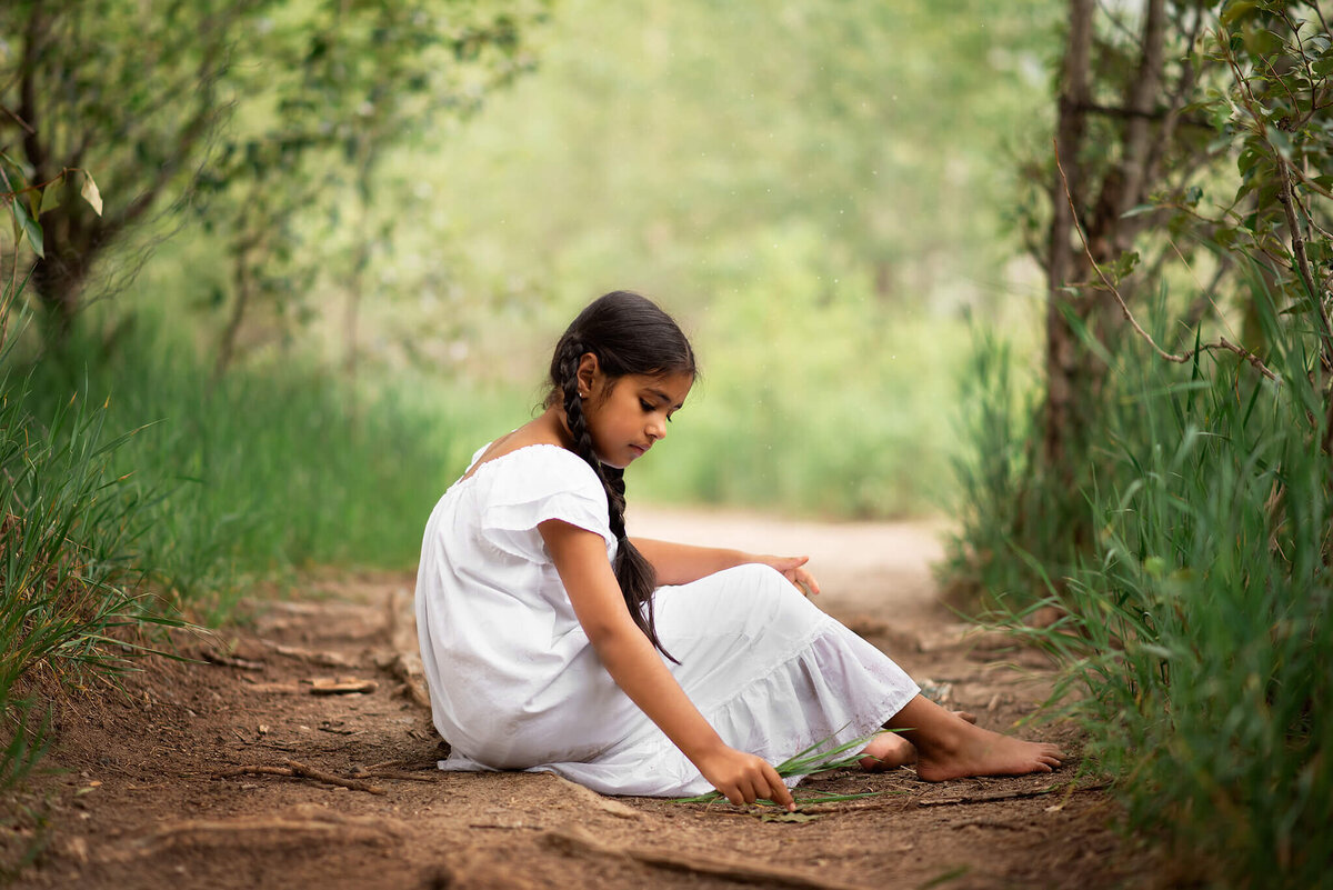 Brittany Danielle photography stuns with this portrait of a young girl with long black hair in braids. She sits on the ground in a white dress amongst green trees.