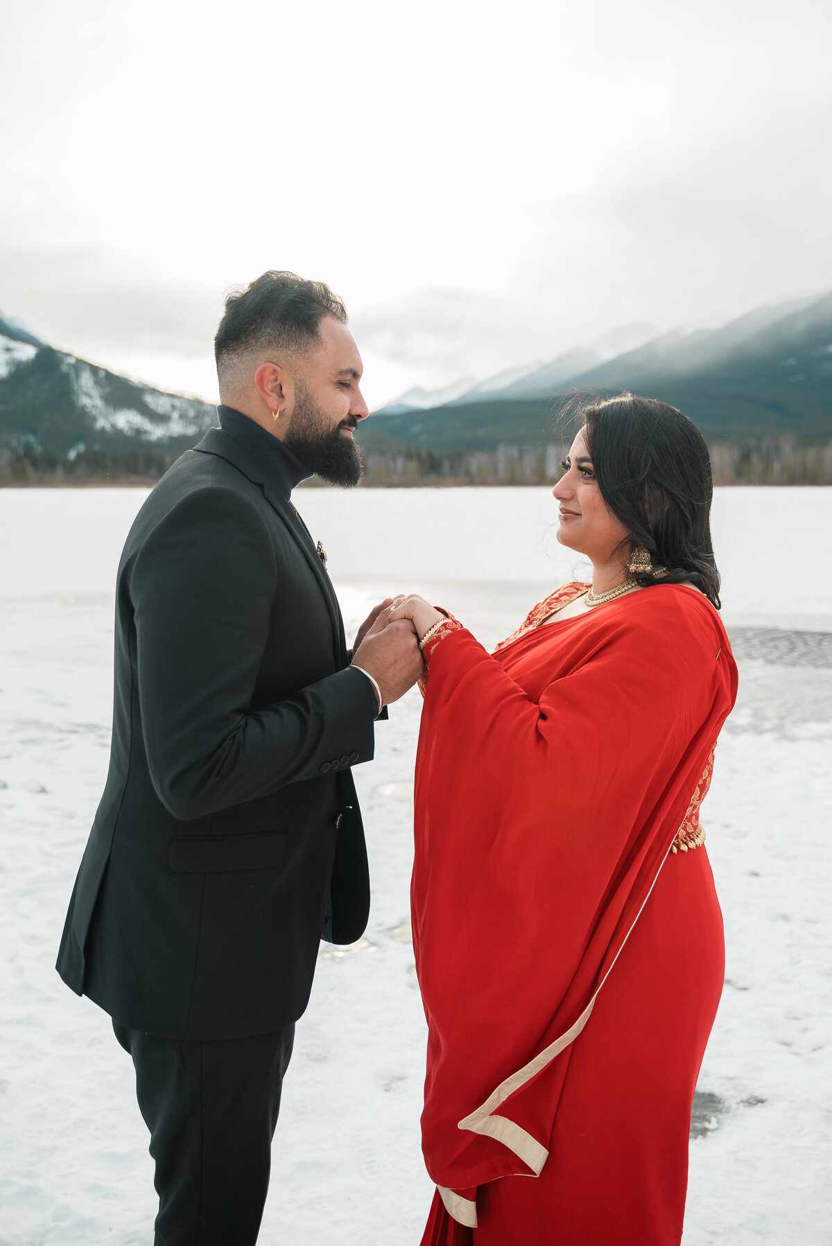 Couple holds hands on snowy dock with Banff mountain reflections.