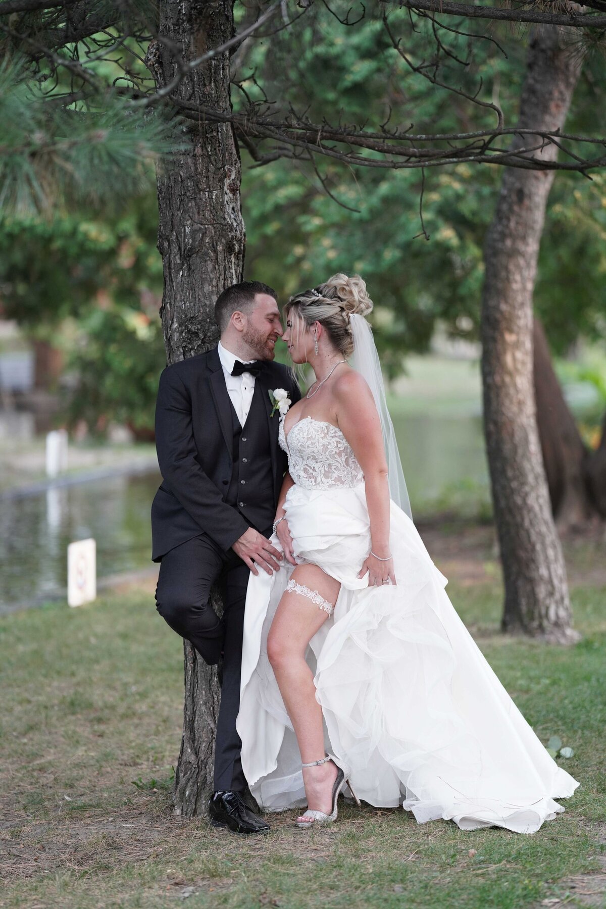 A couple leans against a tree in a playful pose, with the bride proudly showing her garter. The natural setting and their affectionate interaction add a touch of whimsy and romance to the scene.