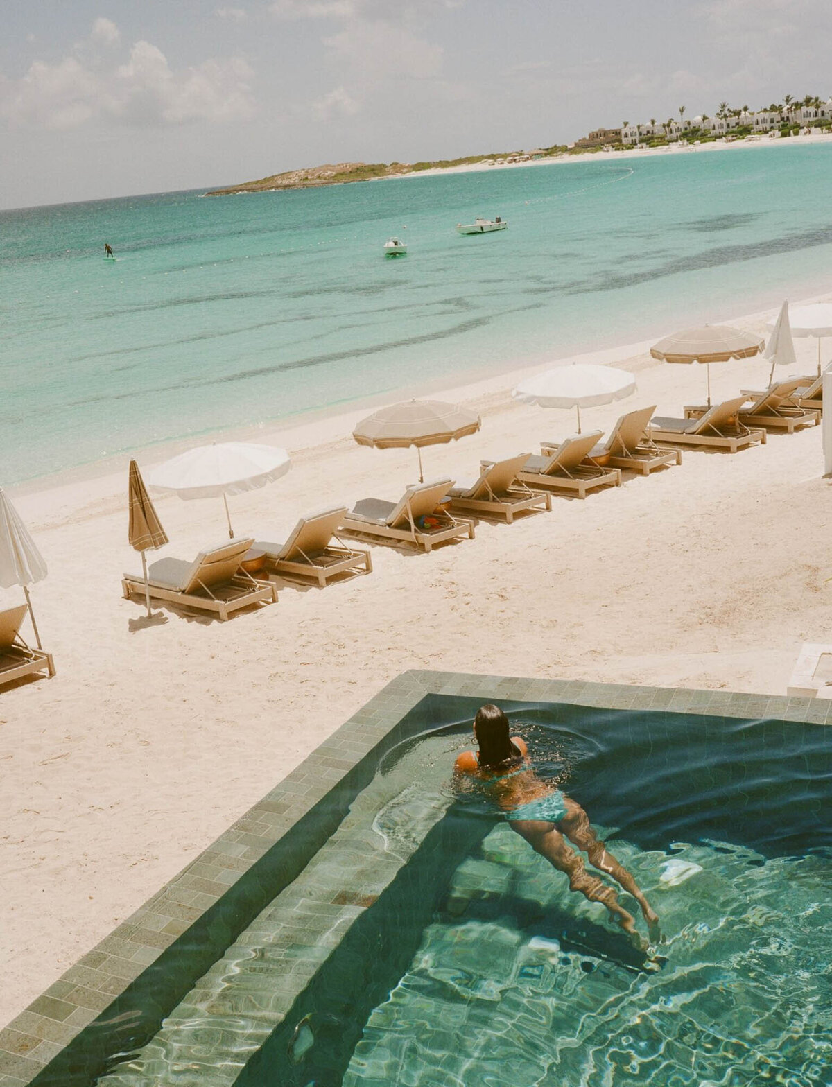 A person relaxes in a pool overlooking a sandy beach with turquoise waters. Empty loungers and umbrellas line the shore under a partly cloudy sky. Paddleboarders can be seen in the distance on the calm sea.