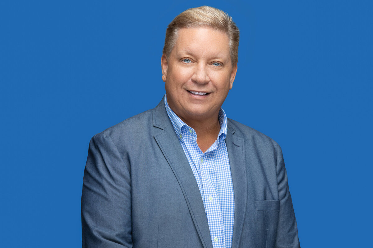 A white businessman in a blue blazer poses for a professional headshot photo on a blue backdrop for Janel Lee Photography studios in Cincinnati Ohio