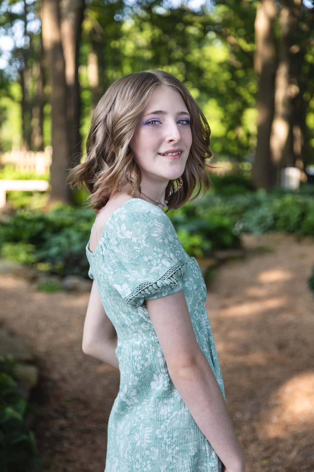 Pretty blonde teen girl wearing a floral green dress looking over her shoulder in the park. Captured by Springfield, MO teen photographer Dynae Levingston.