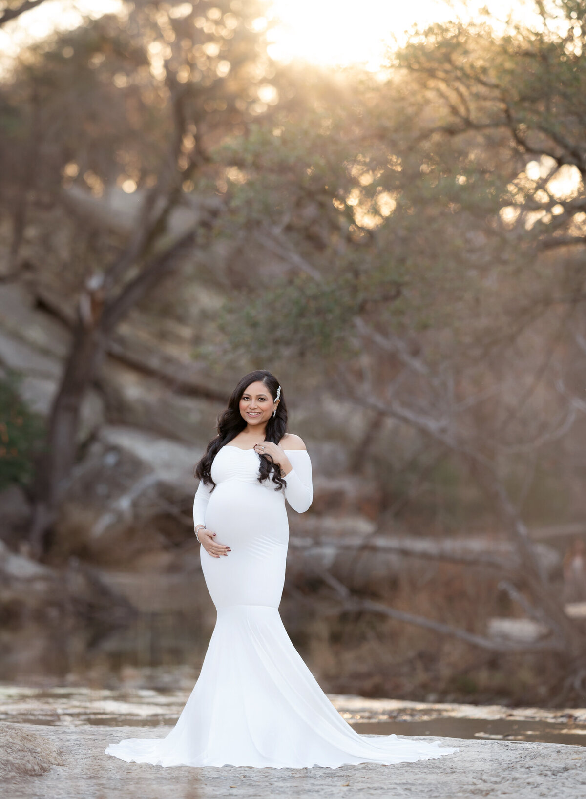 Golden hour maternity portrait in Austin, Texas, with the expecting mom glowing in a long white gown as the sun filters through the trees behind her.