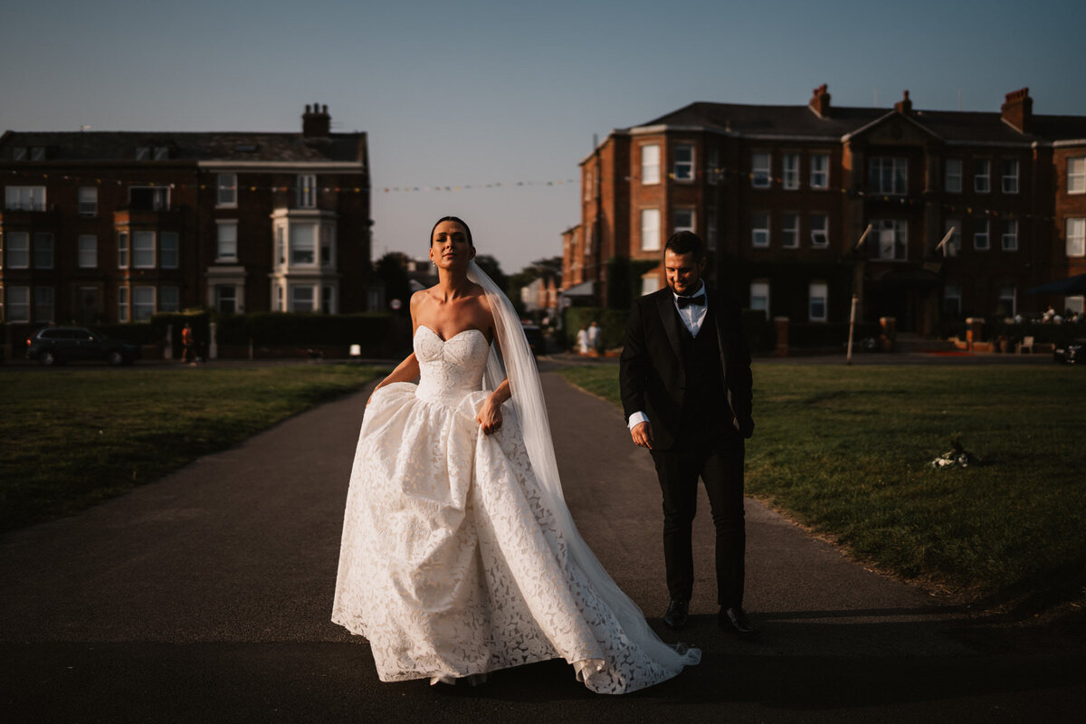 Bride in a strapless lace gown and groom in a black tuxedo walking together at sunset outside a luxury wedding venue in Lytham, capturing an elegant and romantic moment.
