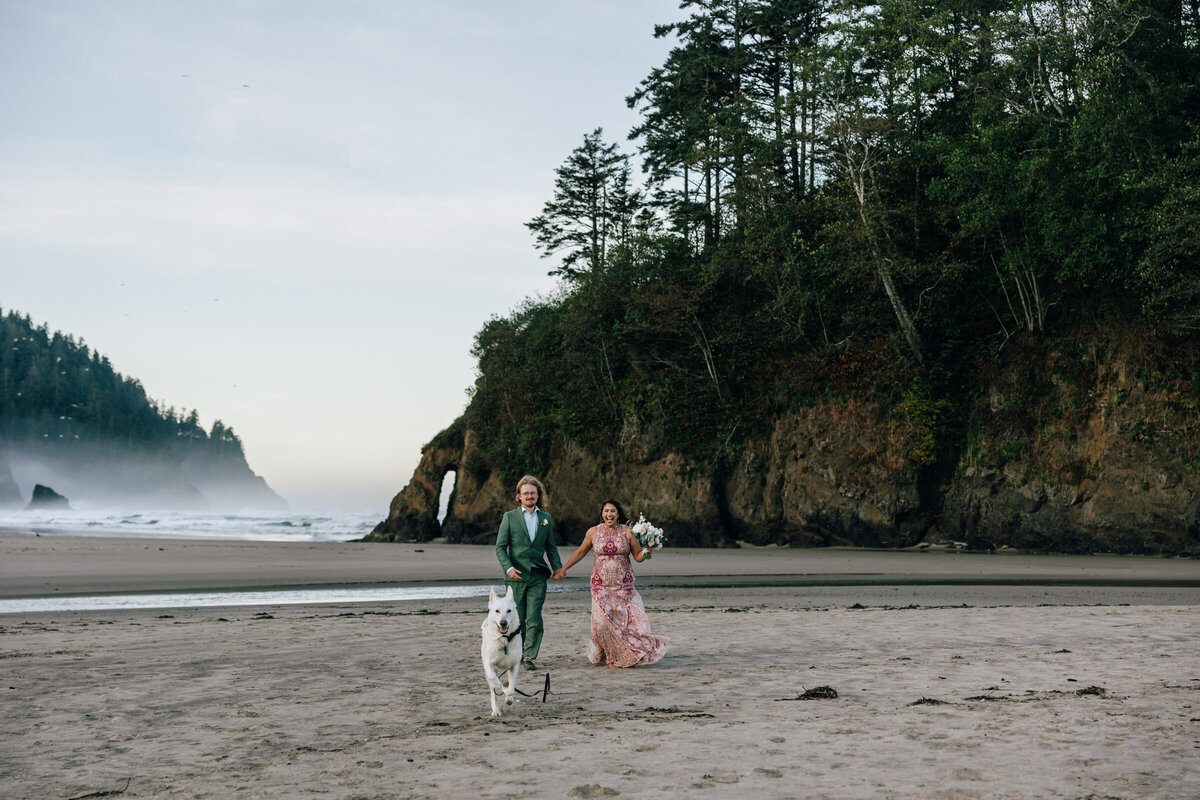 A bride and groom run behind their dog along Neskowin Beach at sunrise during their Oregon Coast elopement. 