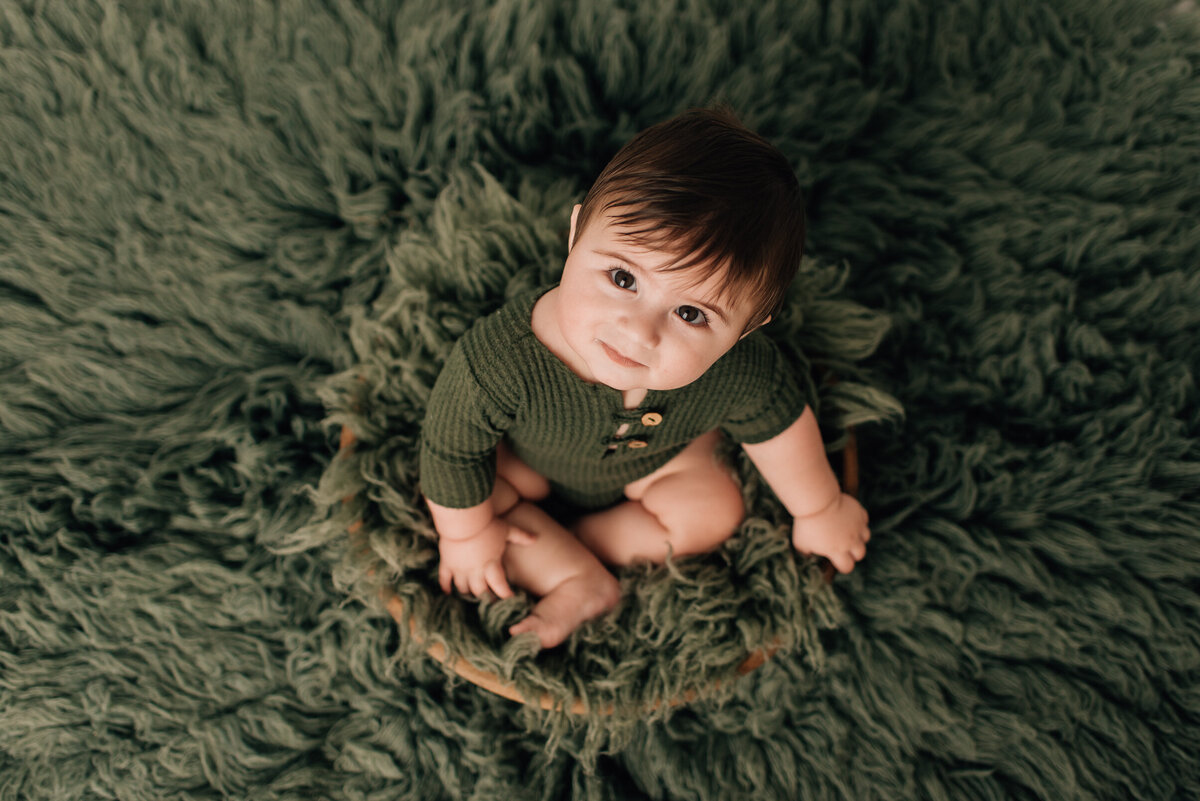 A baby boy sitting on a green rug and looking up at the camera