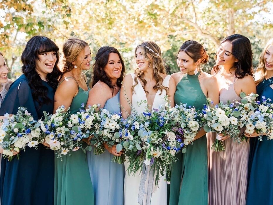  bride in a white wedding gown stands at Palafox Wharf Waterfront Wedding Venue, surrounded by bridesmaids wearing dresses in mauve, light blue, and jade. The bridesmaids hold bouquets of white and blue flowers