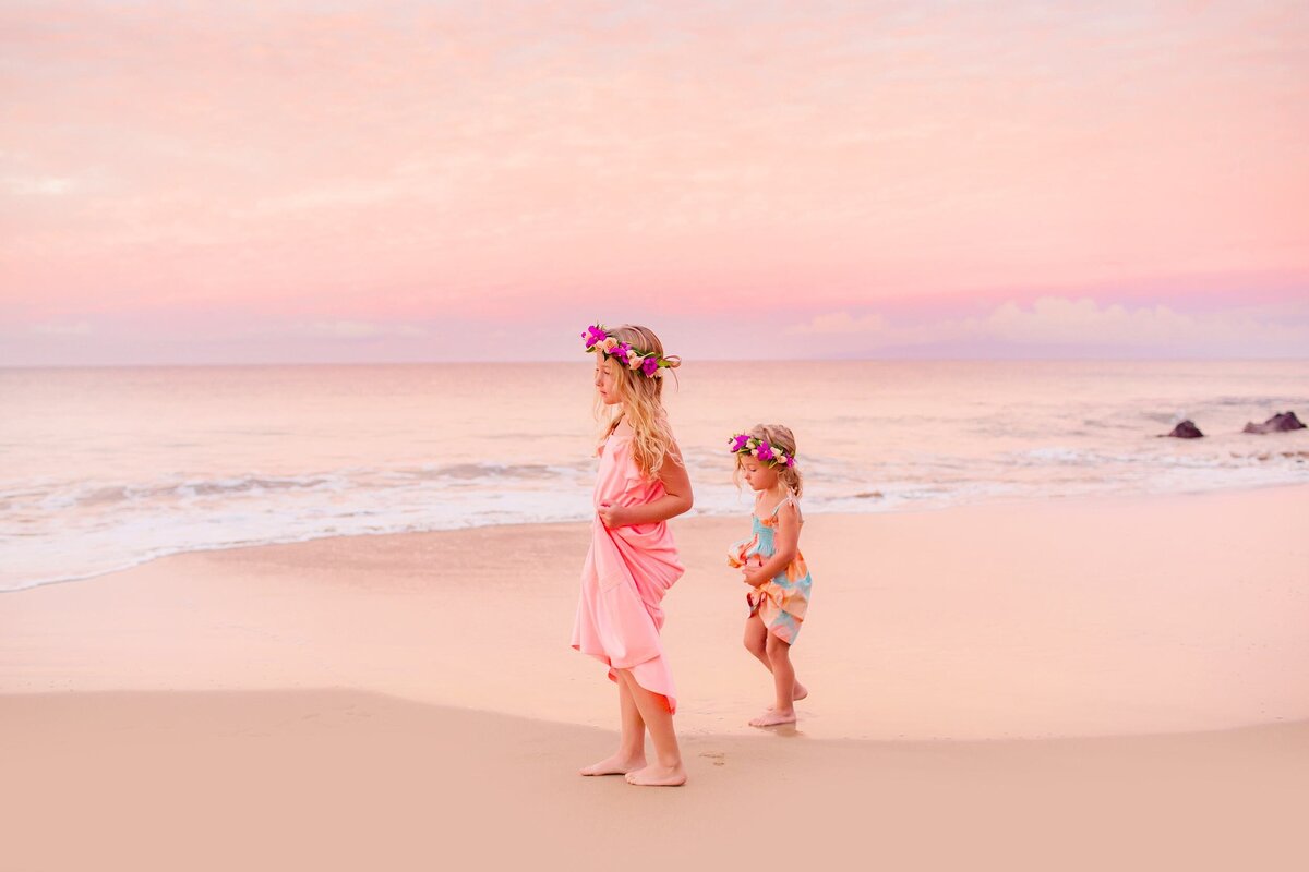 Two young girls wearing flower crowns stand on the sand in Wailea at sunrise with pink sky behind them