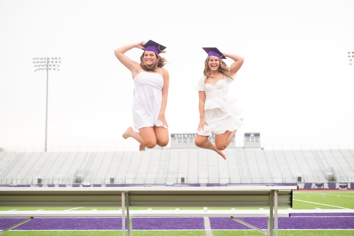 two girls wearing white dressing and purple grad caps jumping up in the air on a football field