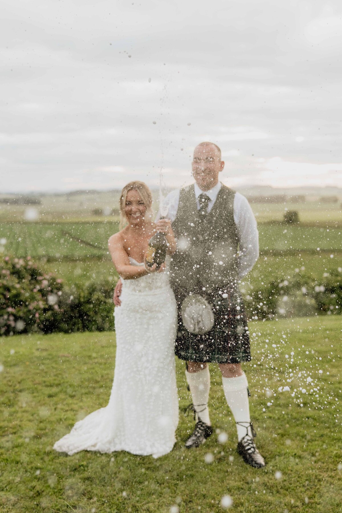 A wide photograph of a bride and groom doing a champagne spray. They wear wedding attire and the Aberdeenshire countryside is seen behind them. The sun sets.