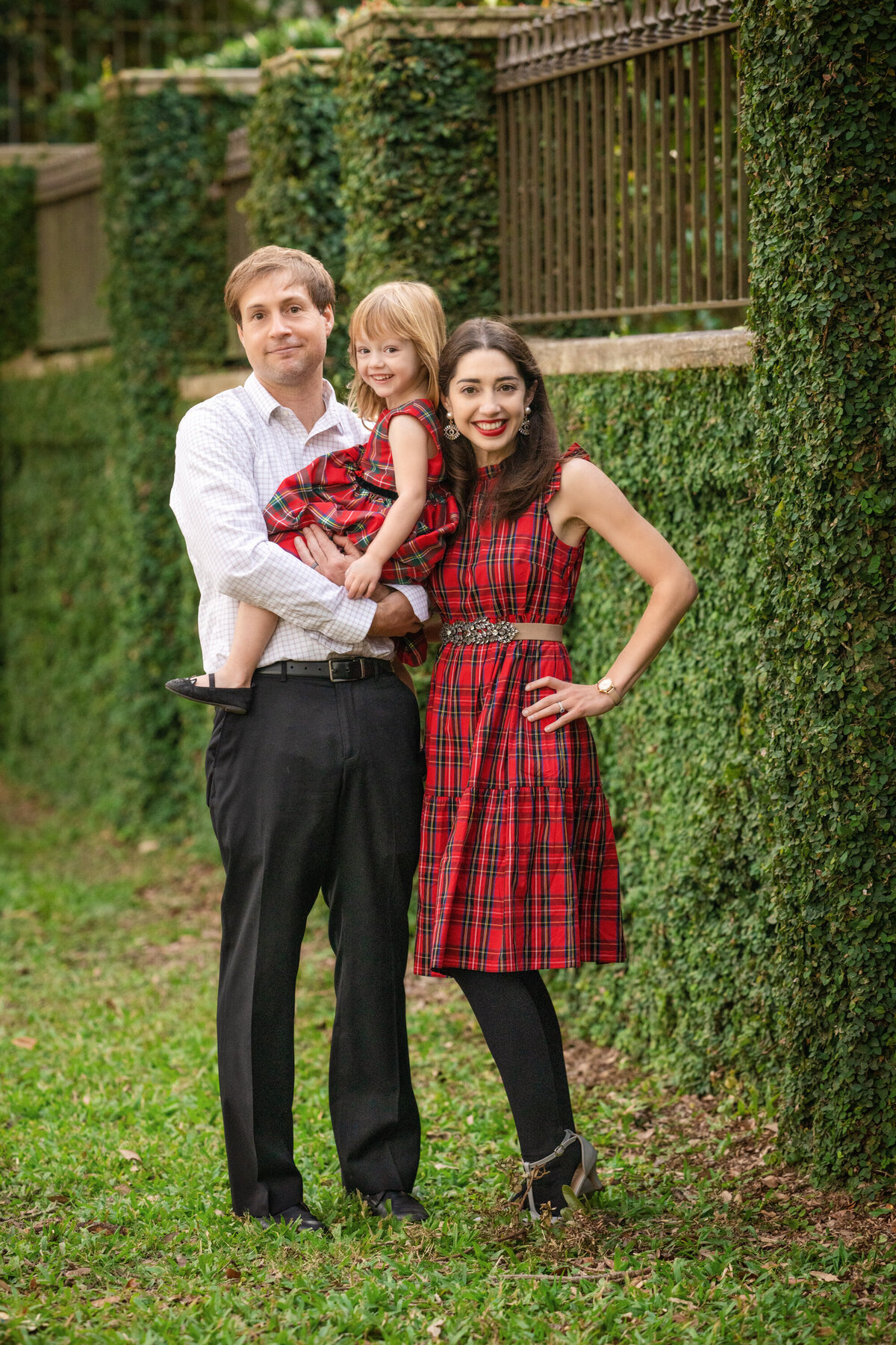Mom and daughter are dressed in red plaid dresses.  Dad is wearing a white dress shirt and black pants.  He is holding the little girl.  They are standing in front of an ivy covered wall.