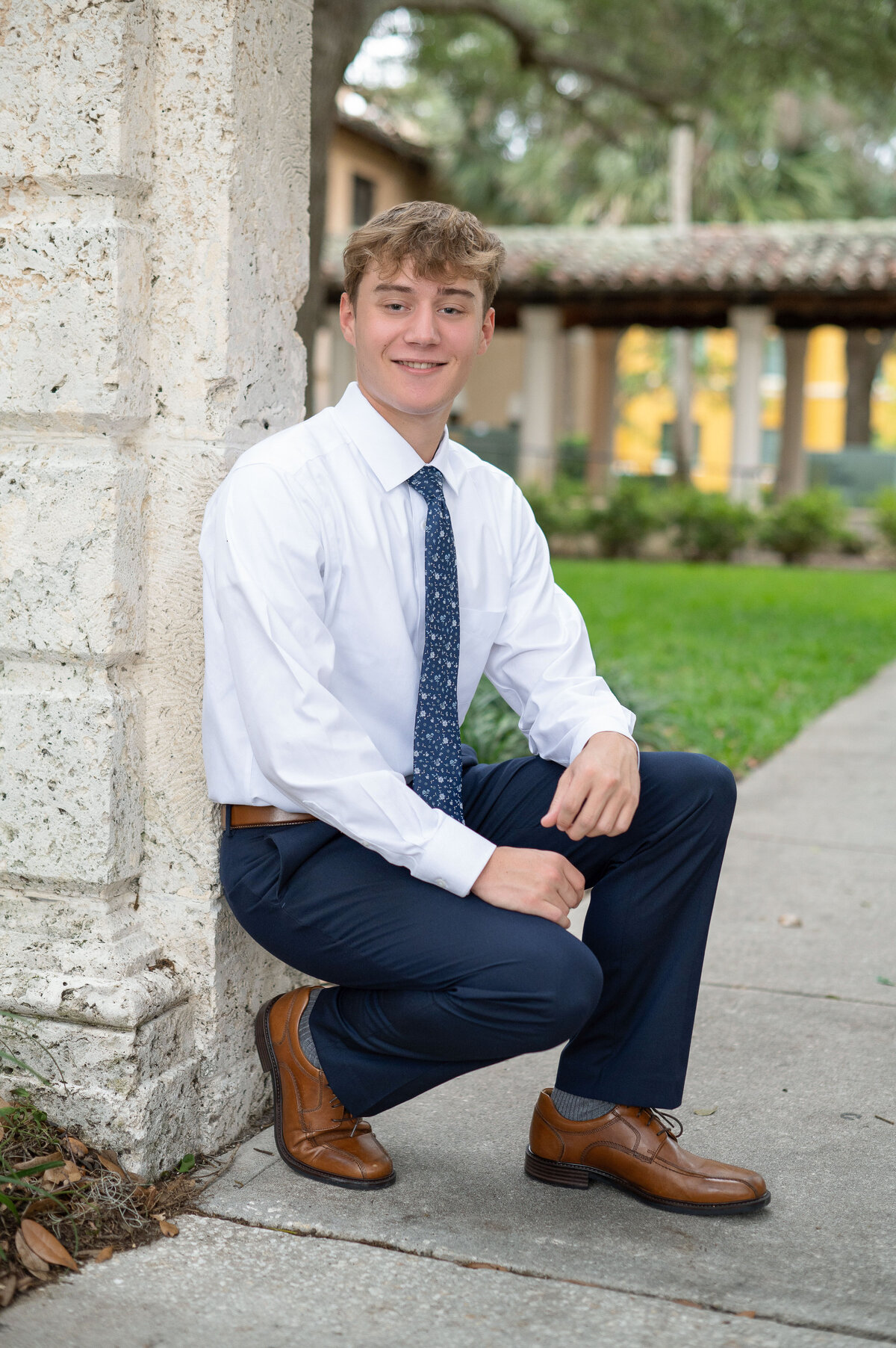 High school senior boy crouching next to a pillar in  a suit tie smiling at the camera.