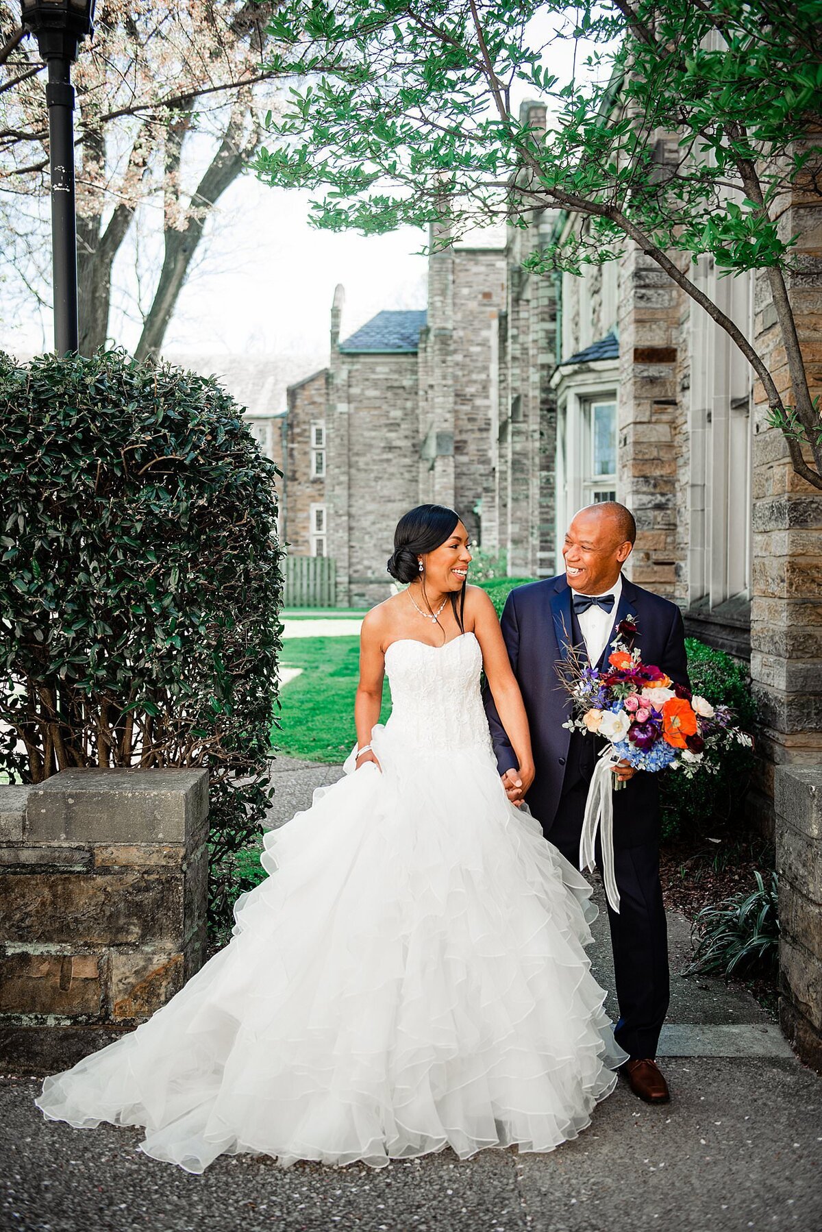 The bride and groom sit outside the large stone church at Scarritt Bennett. The bride is wearing a strapless white wedding dress with a sweetheart neckline on her fitted bodice. The skirt is very full with many layers of ruffles. The bride and groom smile at each other. Th e groom holds the bride's hand as well as her bouquet. The bouquet is made up of deep red, deep purple, yellow, blue lavender, ivory and burgundy flowers with long sheer ribbons accenting the stems. The groom is wearing a black tuxedo with a white shirt and black bow tie.