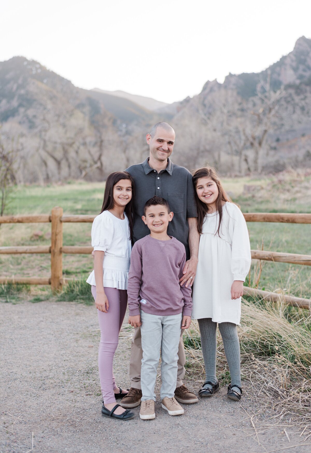 A dad in a grey polo stands with his two daughters and son in front of him while they smile for denver family photographer with the Boulder flat irons behind them captured by denver family photographer