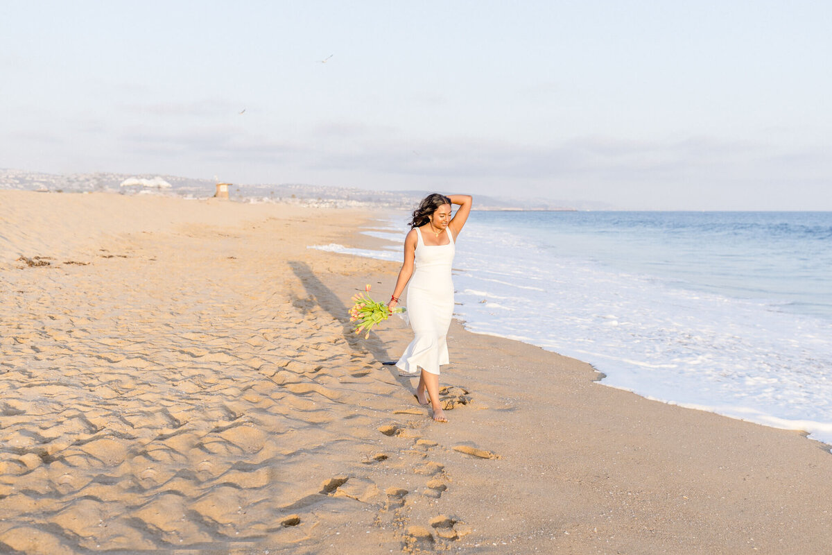 A person in a white dress walks along a sandy beach near the ocean, holding a small bouquet of flowers. The sky is clear and the sun casts a warm glow on the scene. Footprints are visible in the sand.