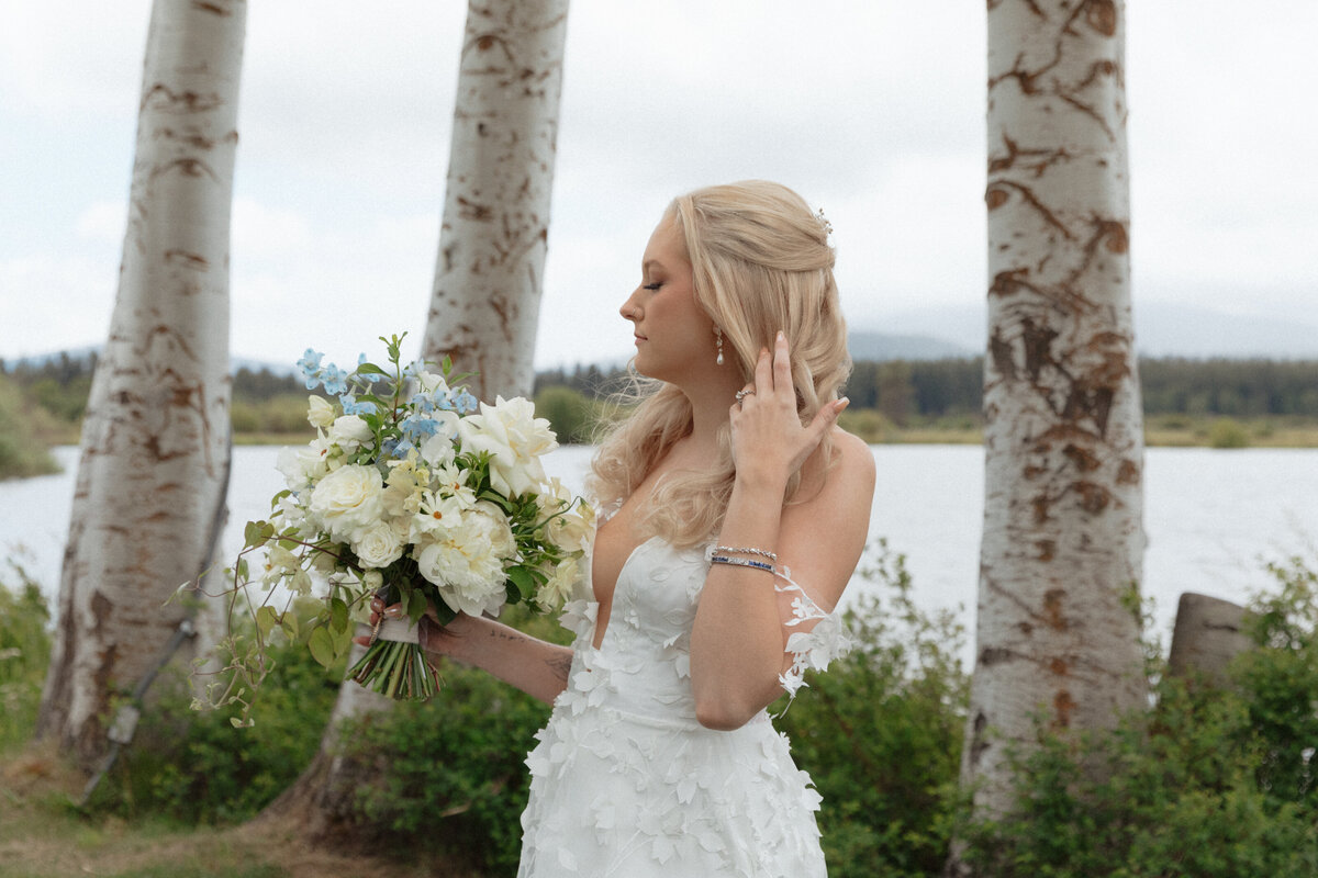 Bride stands in sunlight, holding her bouquet and pushing back her hair in front of three trees.