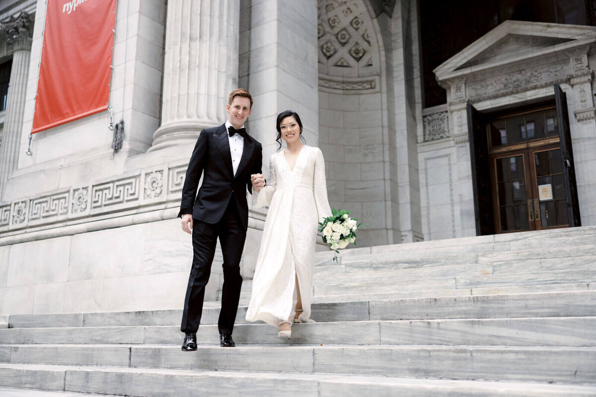 The bride and the groom are on the New York Public Library's grand staircase entrance with large marble columns and walls.