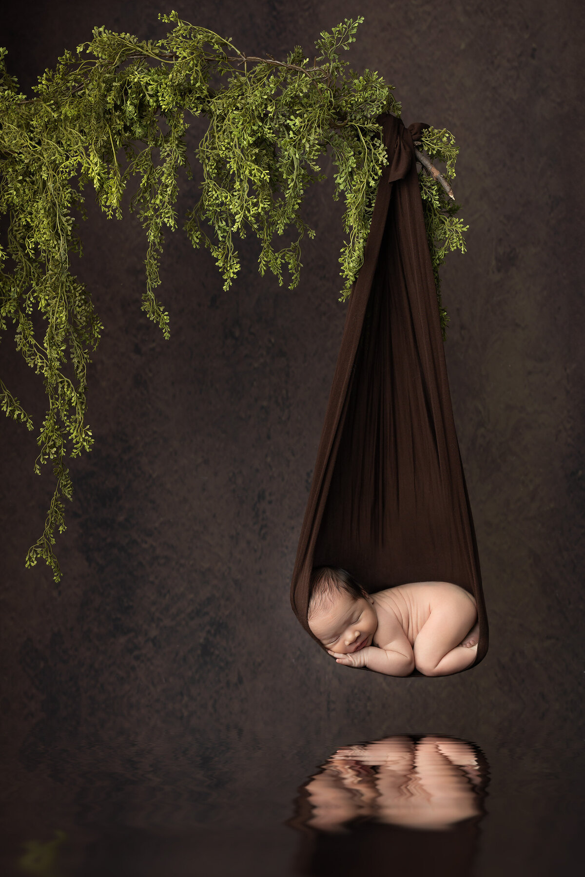 Newborn baby curled up in a brown hanging wrap suspended from greenery, reflected softly below on a water-like surface, photographed by a Dallas newborn photographer in a dreamy, nature-inspired setup.