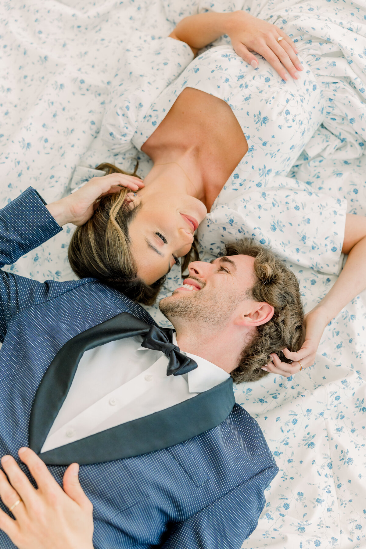 Ivy Rose Barn | A bride in a vintage-inspired puff sleeve floral gown and groom in navy tuxedo lay on the train of the bride’s dress. The pair cup each others’ faces as they lay in the fabric.