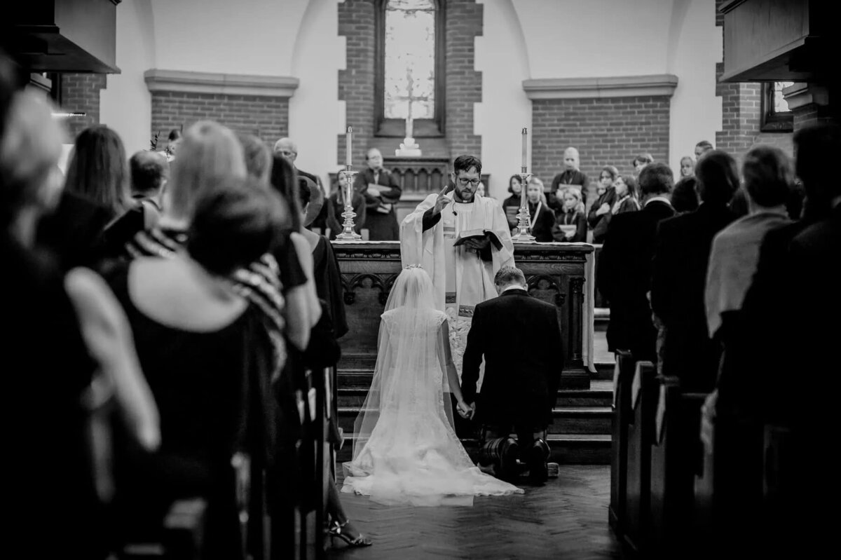 A bride and groom kneeling in front of a priest during their wedding.