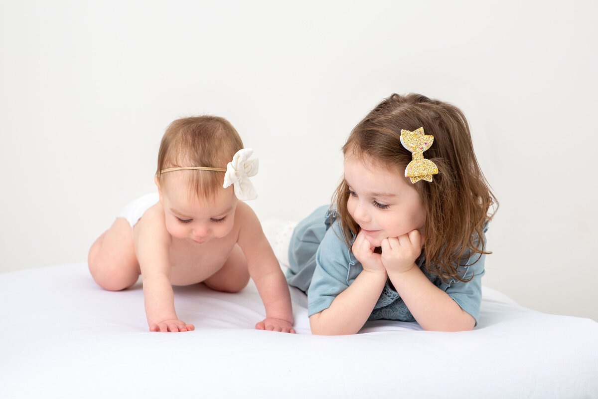 Young toddler girl and her baby sister lying on their bellies during a photo session, sharing a tender moment with curious expressions and close sibling connection