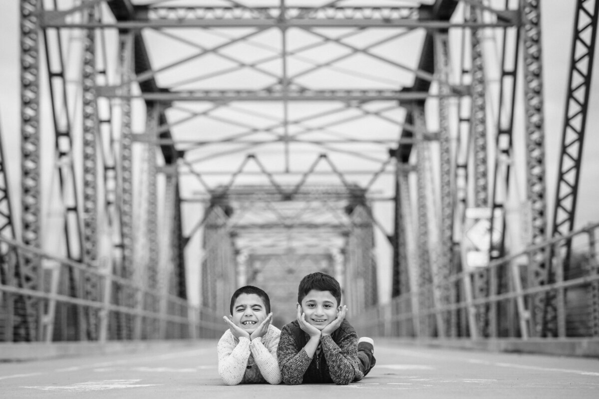 Two boys lying on their stomachs, side by side, on a metal bridge, resting their chins on their hands.