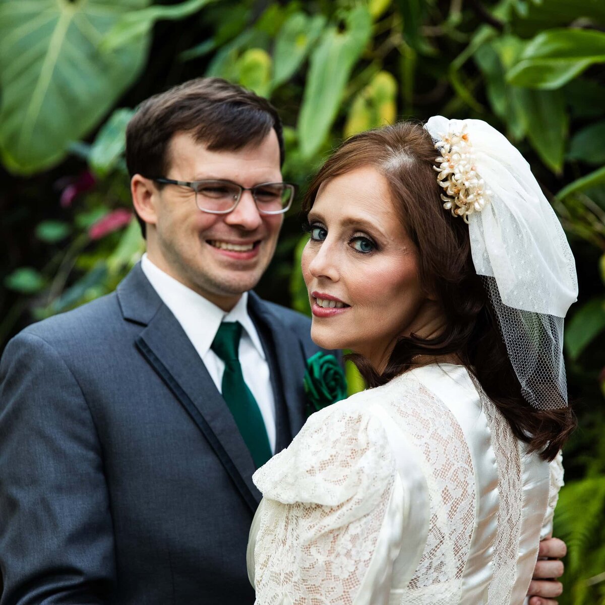 Bride and groom in a lush tropical environment at an Aquarium wedding, composed close to the subject. The bride is glancing over her shoulder at the camera while the groom looks at her and smiles