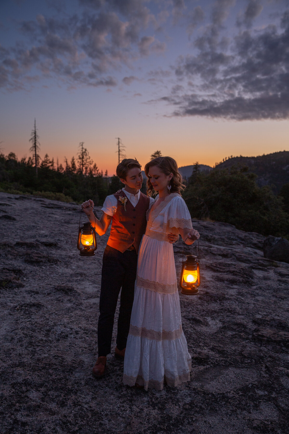 Two brides stand hugging each other while holding lanterns next to their bodies as the sunsets in Yosemite.