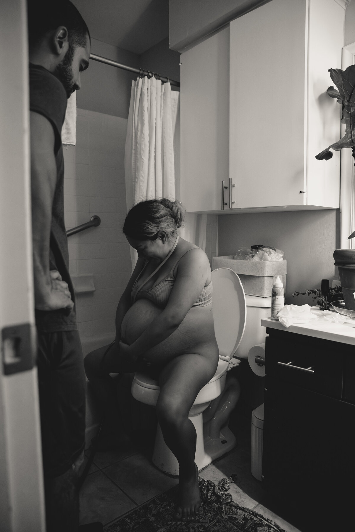 Mother labors down through contractions on the toilet, husband is onlooking and supporting from nearby