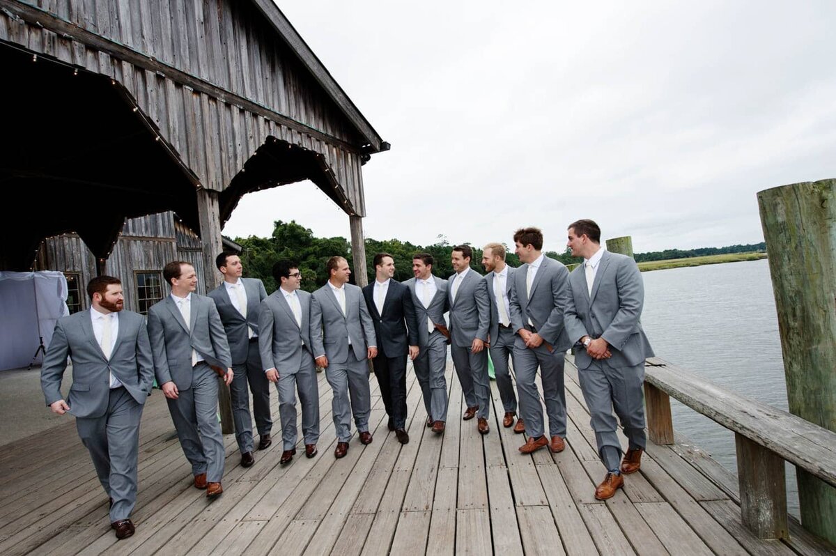 a large crew of groomsmen walk along a dock on the river