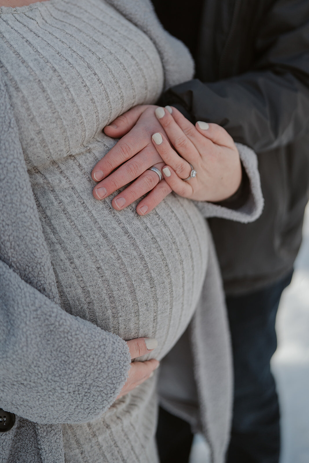 Close-up of a pregnant belly with both parents’ hands resting on it during a snowy maternity session at Al Sabo Preserve in Kalamazoo Michigan.