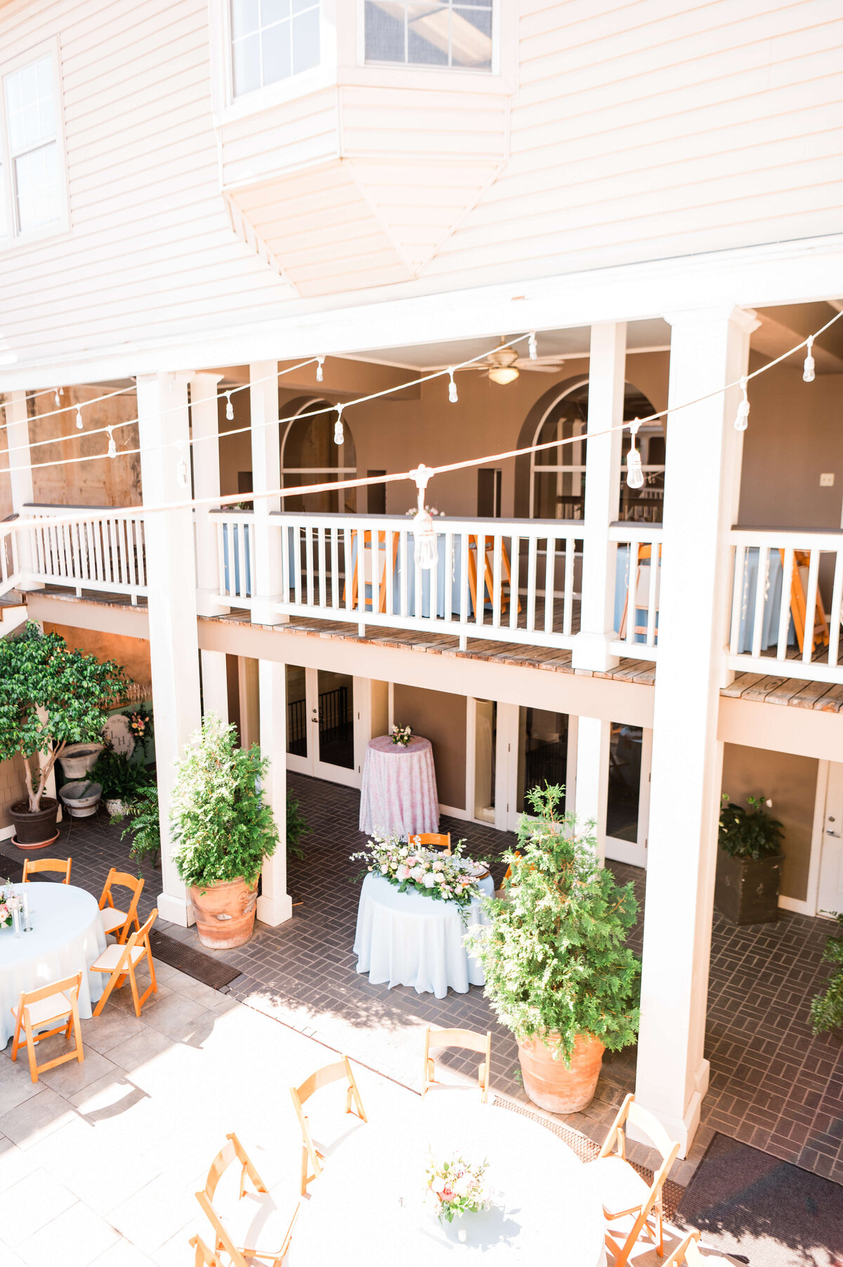 A bright courtyard set up for an event features round tables with light blue tablecloths, wooden chairs, potted plants, and string lights hanging above, bordered by two-story balconies.