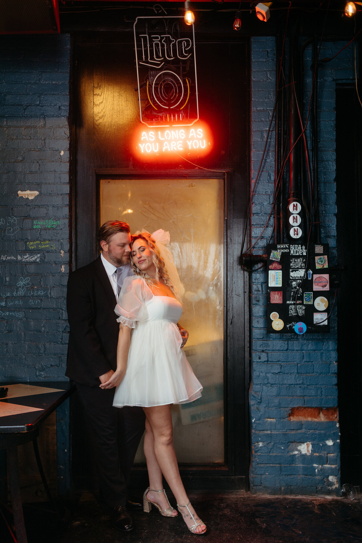 An affectionate couple stands in front of a vibrant neon sign reading 'Live as long as you are you'