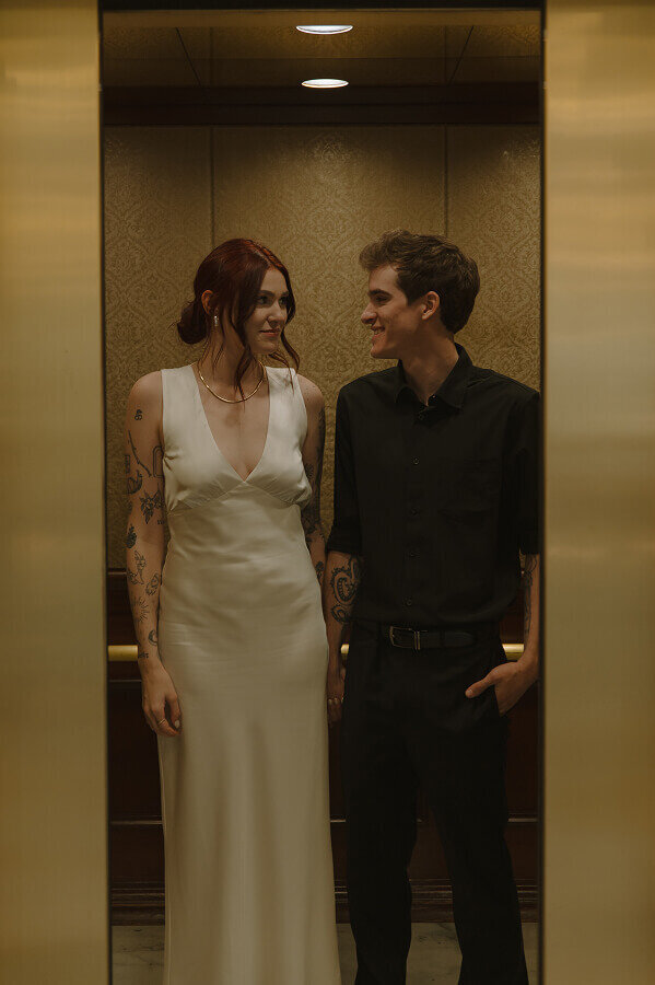 Standing in an elevator, a man and woman stand side by side looking at each other.