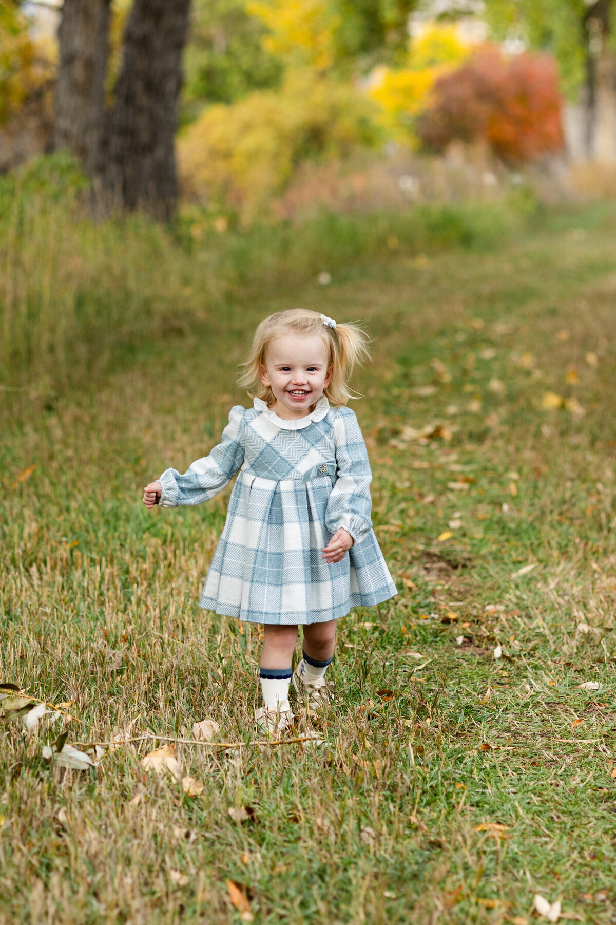 Toddler girl in a blue and white plaid dress walks toward the camera on a wooded path and smiles.