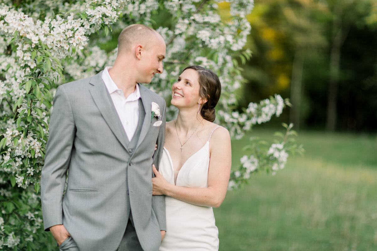 A bride smiling at her groom  and holding his arm in front of a flowering apple tree in Door County, WI