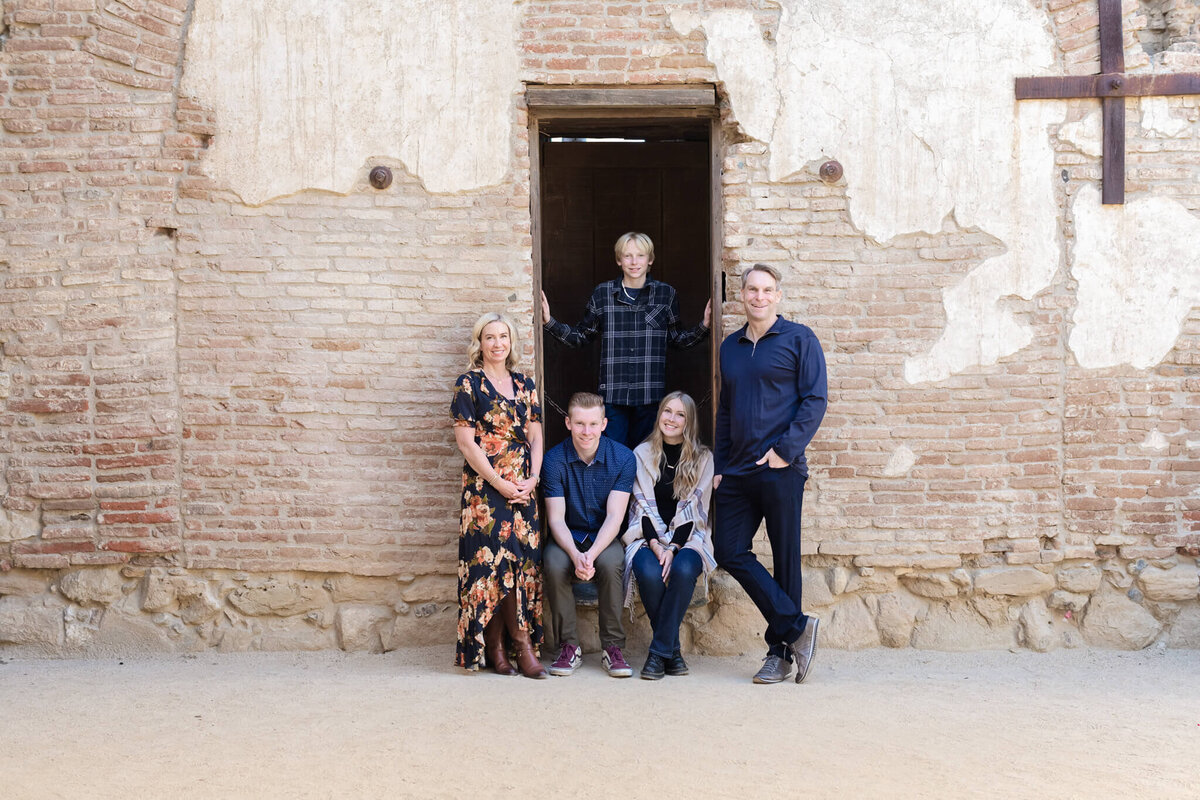 A family of five sitting and standing around an old wooden doorframe in an old beige brick wall. Captured far away at Mission San Juan Capistrano.