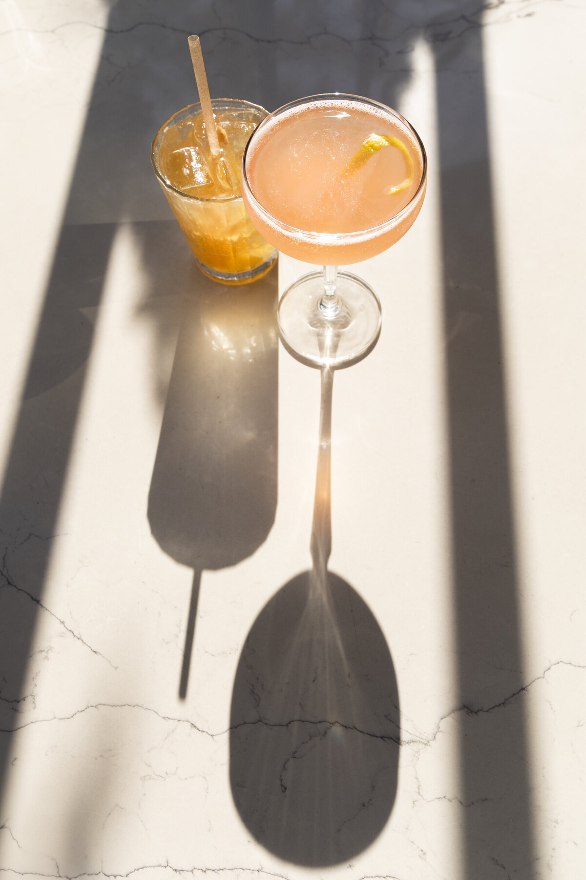 A dark and stormy and a pear flower cocktail  with long shadows and light refractions on a marble table.