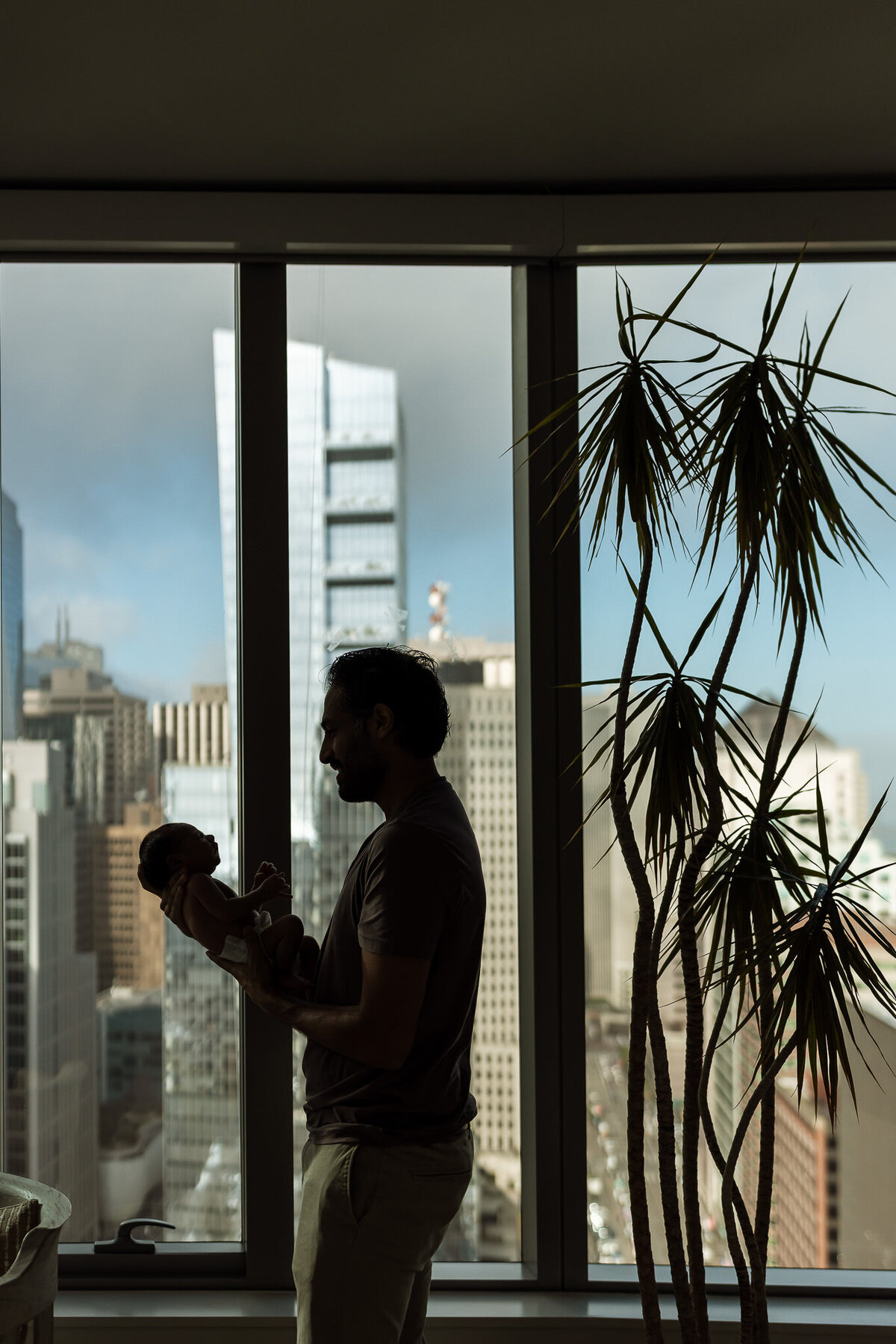 In home newborn being held by father in front of a window with San Francisco skyline as backdrop